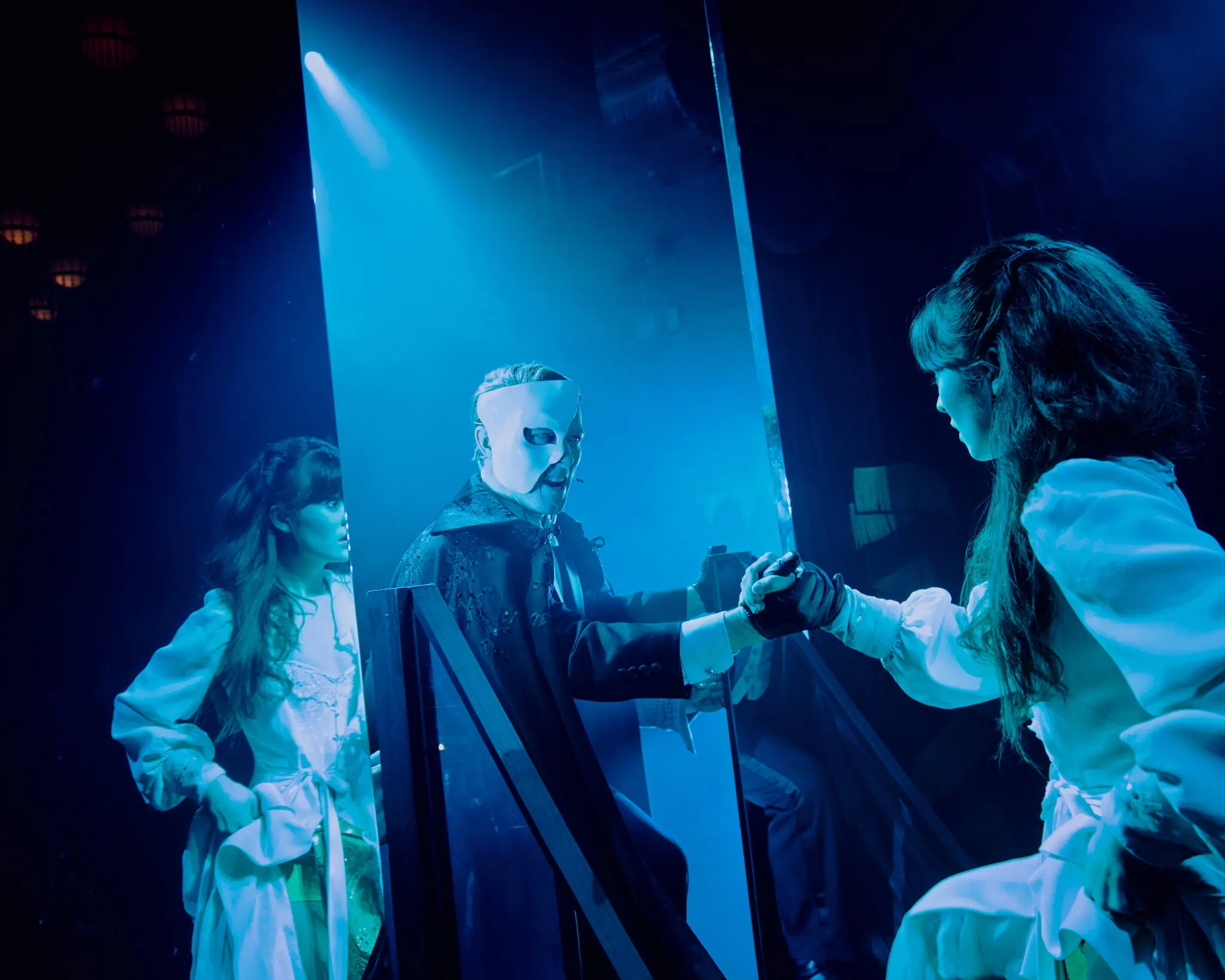 The Phantom in a white half-mask and dark caped coat steps through a tall vertical mirror, reaching out to take the hand of Christine in a white period dress, while her mirror-image reflection is visible on the left side of the glass. Cool blue stage haze fills the background.