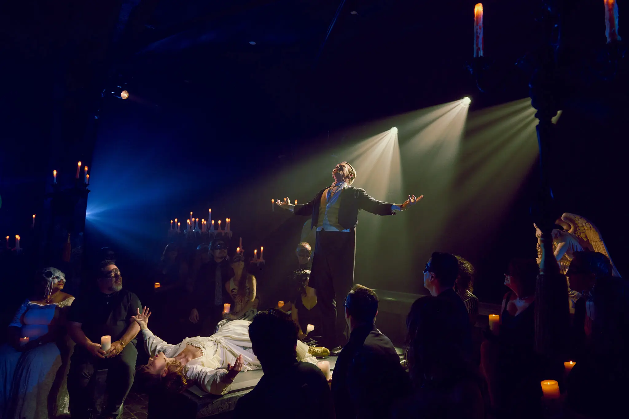 The Phantom of the Opera stands center stage with arms outstretched, lit by a tight overhead spotlight with visible light rays cutting through haze, while Christine in a white gown lies prone on a platform below him. Masked audience members holding candles press in on all sides, with candleabras glowing in the background.