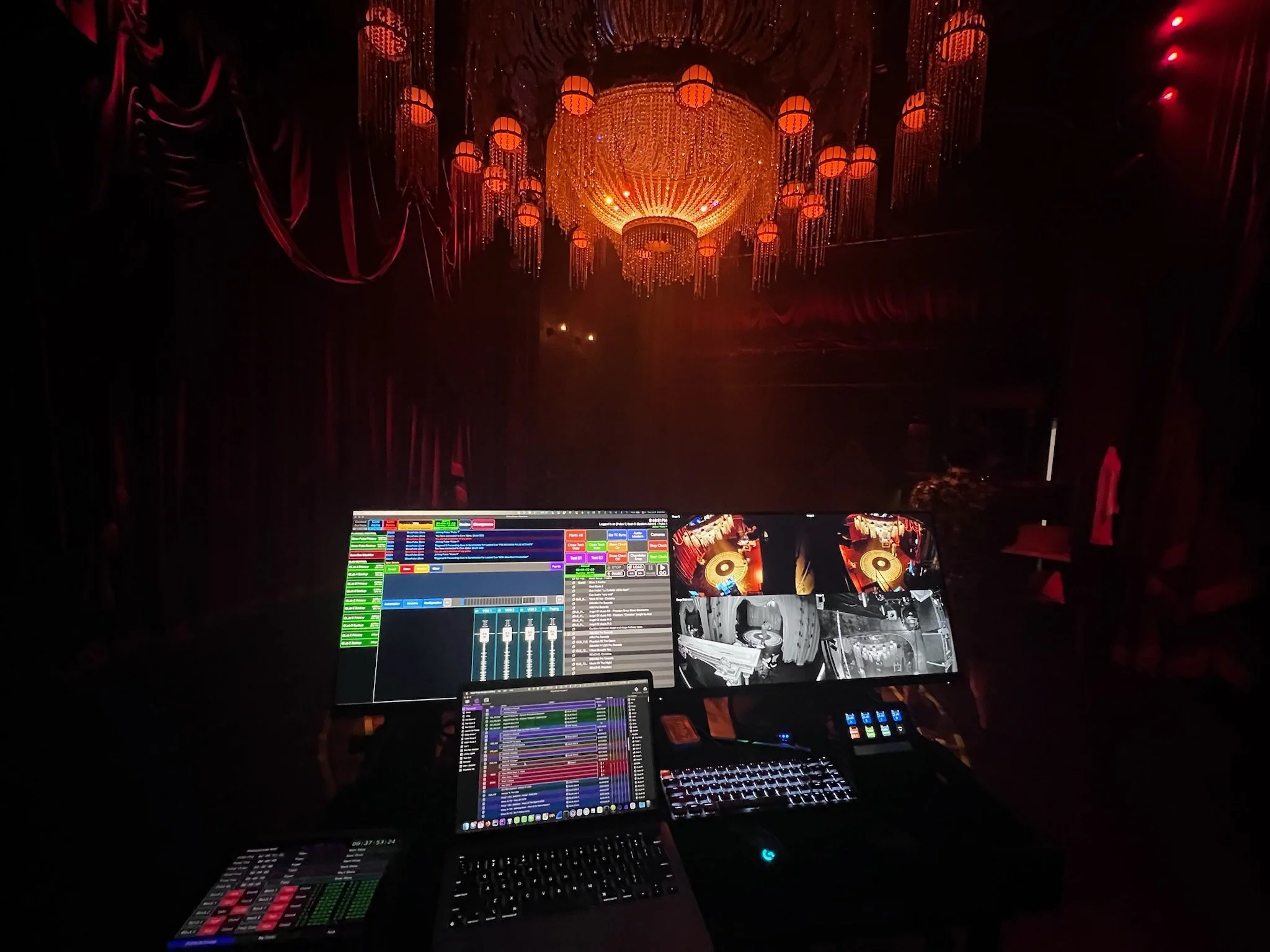 The ShowPulse operator workstation photographed from behind during a live Masquerade performance, with three screens visible: the ShowPulse runtime interface on the left monitor, a multi-camera feed showing live views of the chandelier and performance spaces on the right monitor, and a laptop running additional show control software in the center. The production's massive crystal chandelier glows amber overhead in the darkened performance space beyond the screens.