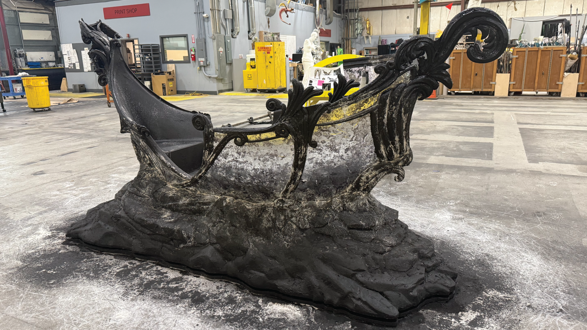 A large ornate gondola stage prop photographed on the floor of a scenic fabrication workshop. The piece is painted matte black with weathered gray highlights and features elaborate baroque scrollwork along its hull and prow, mounted on a sculpted base textured to resemble dark stone or water-worn rock. Industrial shop equipment, crates, and a flammable materials cabinet are visible in the background.
