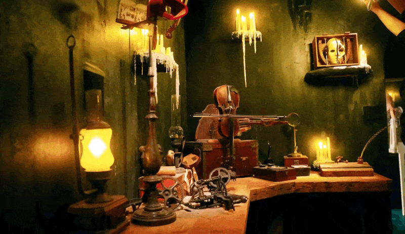 The Phantom's underground lair set, featuring a cluttered workbench with antique mechanical gear assemblies, a violin resting on a travel trunk, dripping wax candelabras, and an oil lamp casting warm light against moss-green distressed walls.