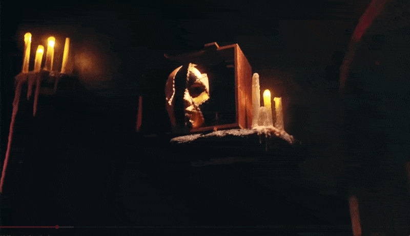 A white Phantom half-mask, cracked and torn at its edges, emerging from a small wooden box surrounded by melted wax candle stumps, dramatically backlit against a near-black background.