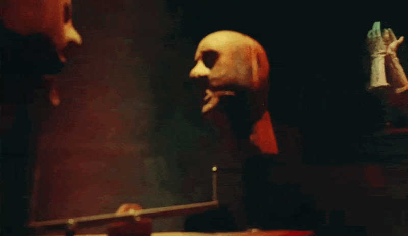 Two full-face theatrical masks on stands facing each other in near-darkness, one warm-toned and rounded, the other lighter with a more angular profile, lit by faint candlelight in the background.