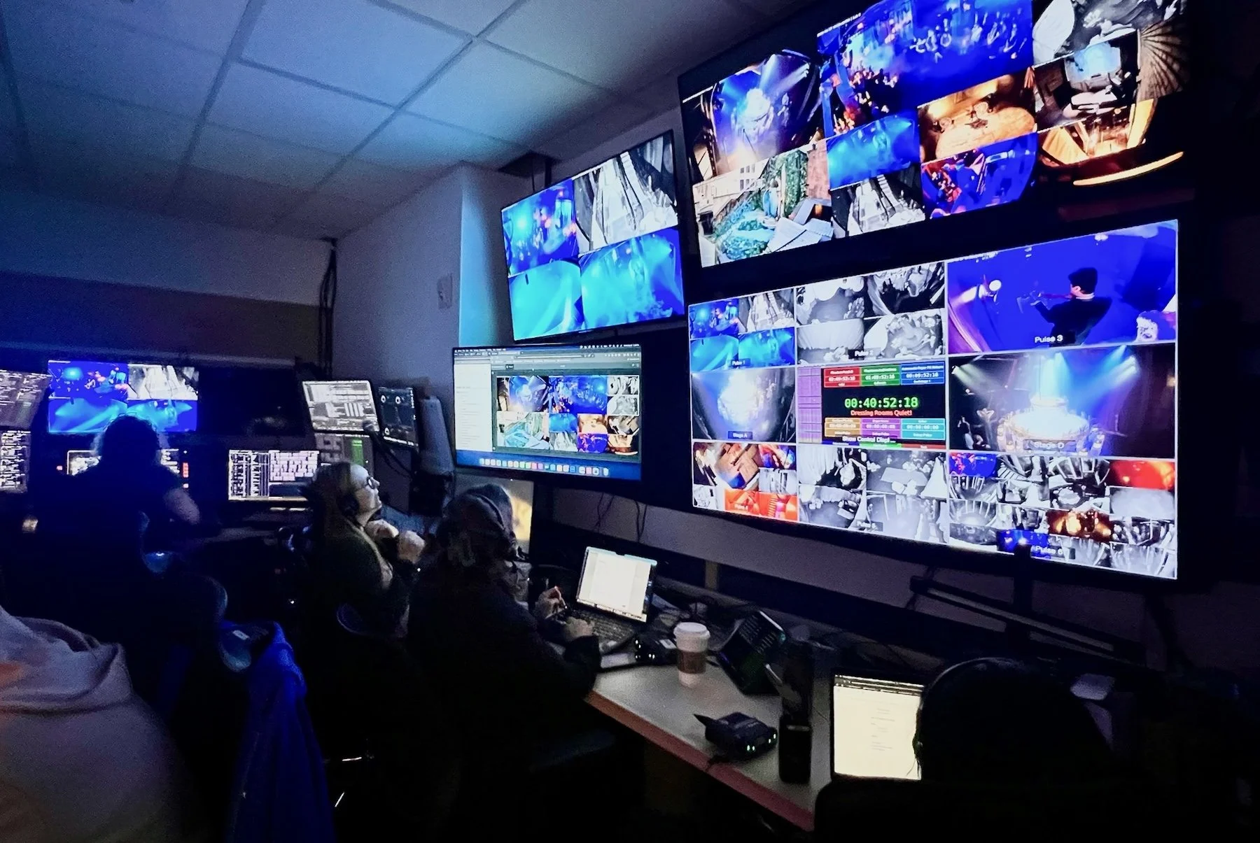 The Masquerade production control room during a live performance, showing three crew members at workstations facing a wall of large monitors. The screens display a multi-camera grid of live feeds from performance spaces throughout the building — labeled by pulse number — alongside a large timecode display reading 00:40:52:18 with the cue annotation "Crossing Rooms Quietly" visible in green text. Additional monitors show show control software interfaces and camera switching views.